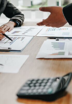 Foto ilustrativa de relatórios em cima de uma mesa, com duas pessoas conversando ao redor.